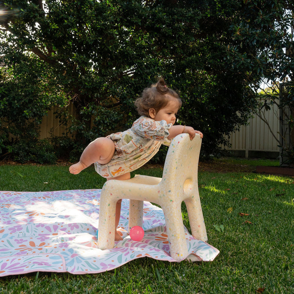 Child playing on a chair outdoors with pink balls and a colorful blanket.
